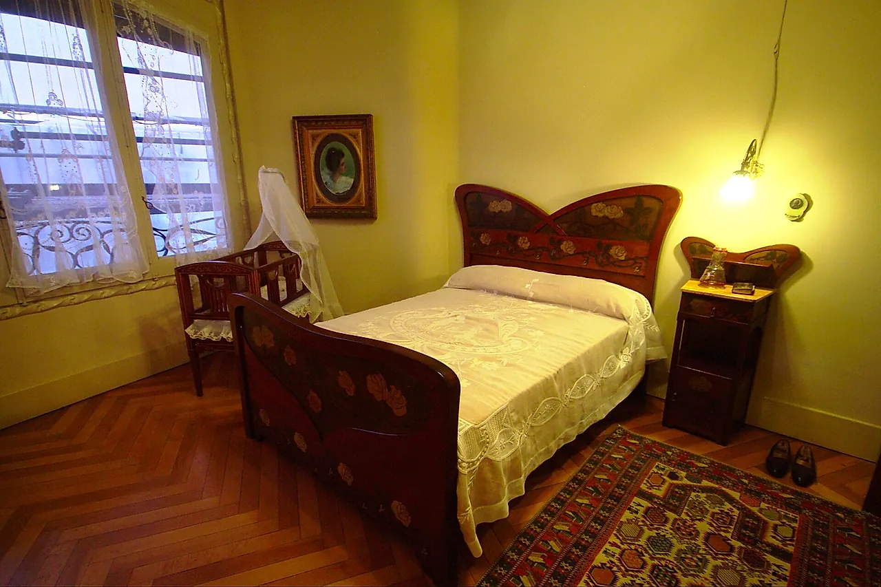 A bedroom of the period apartment in La Pedrera, with a carved wooden bed and bedside table, a parquet floor and a patterned rug
