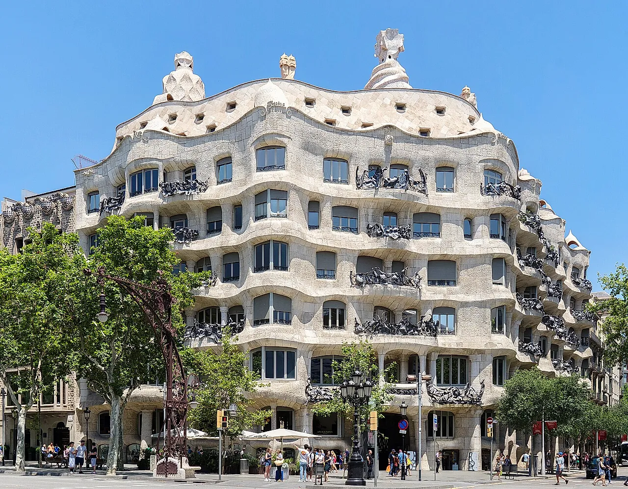 The undulating stone facade of Casa Milà seen from Passeig de Gràcia on a clear day, with its wrought-iron balconies and the plane trees along the pavement